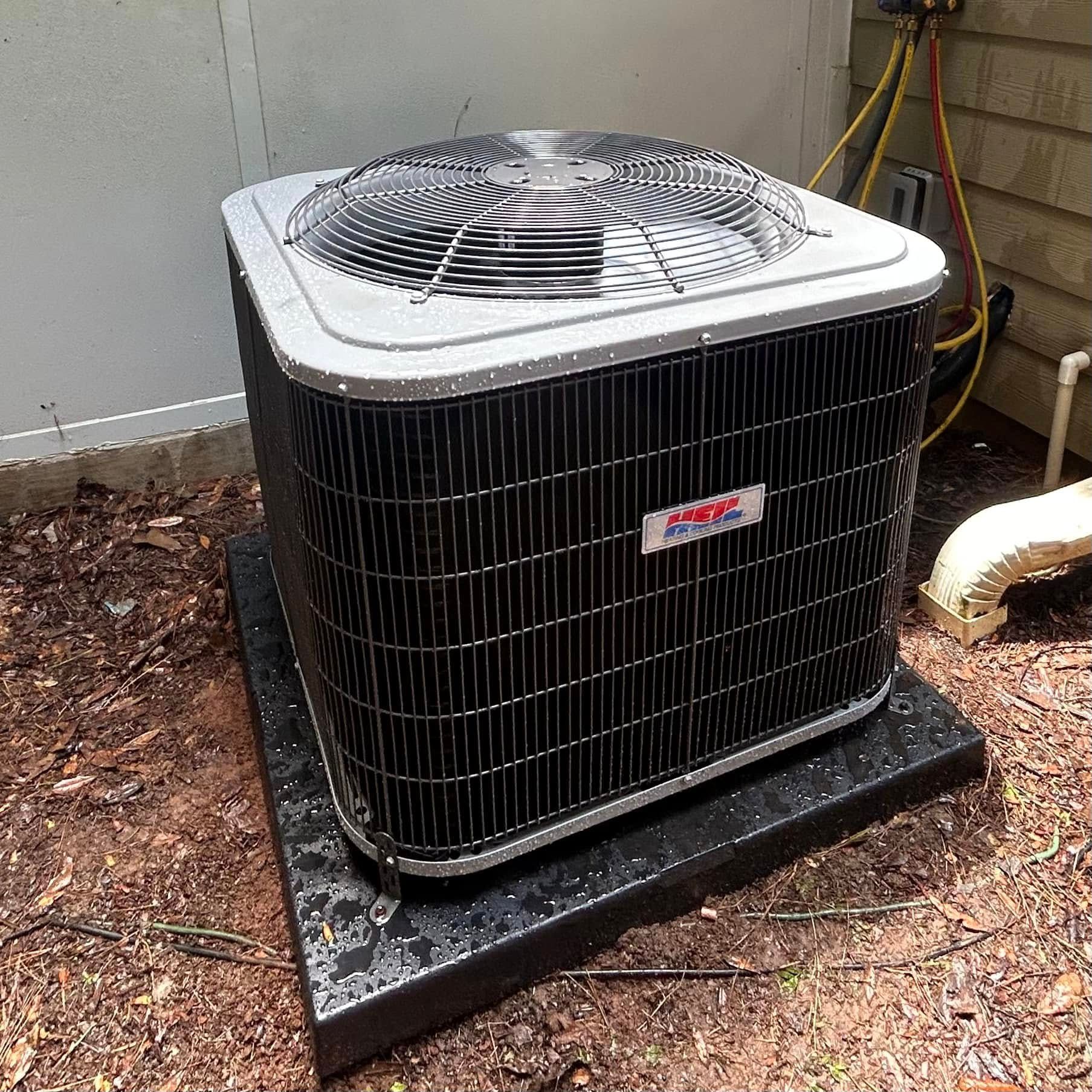 Outdoor air conditioner unit on a black pad beside a wall, with a fan grill on top.