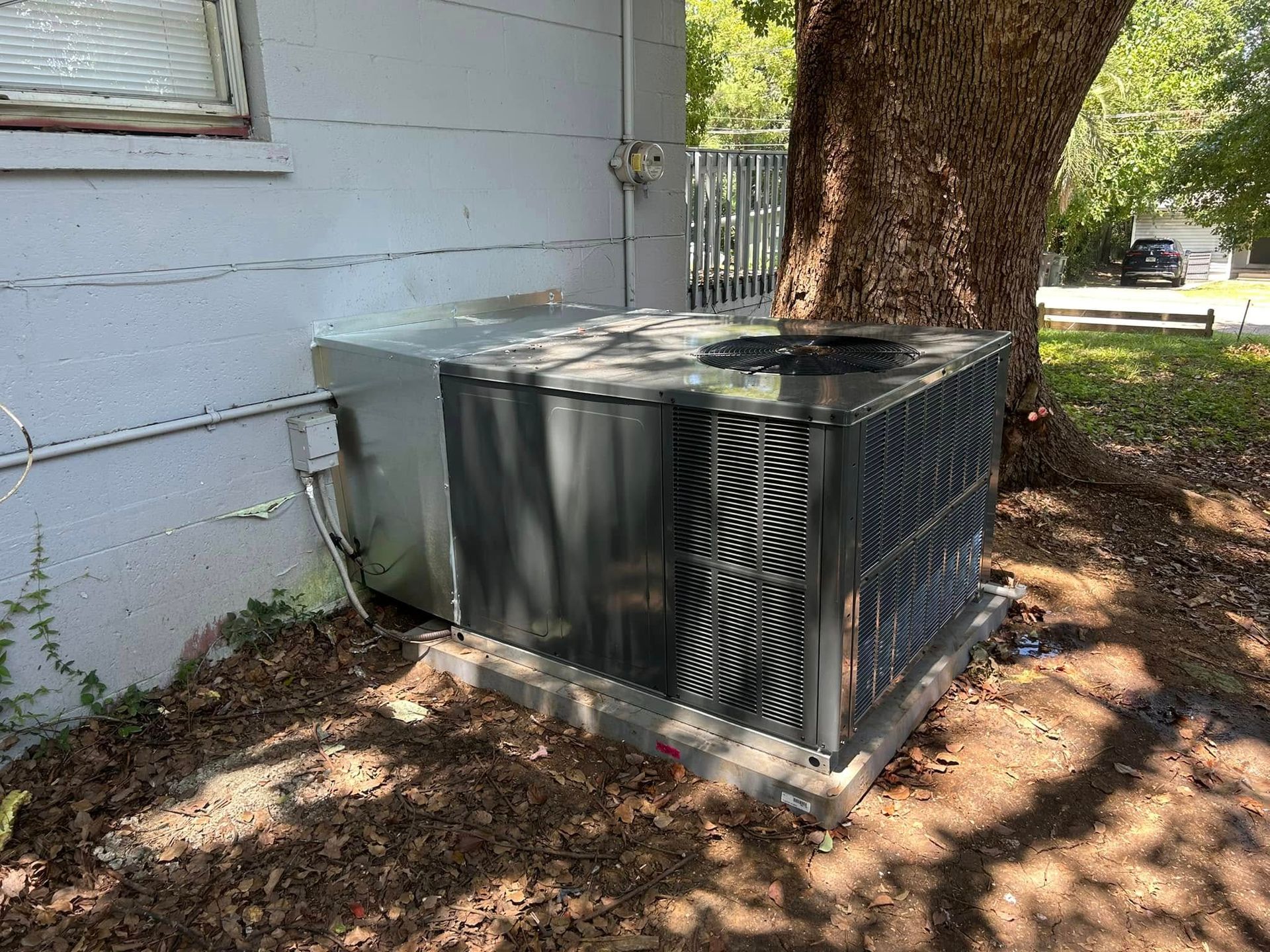 Outdoor air conditioner unit beside a house and large tree, with leaves scattered on the ground.