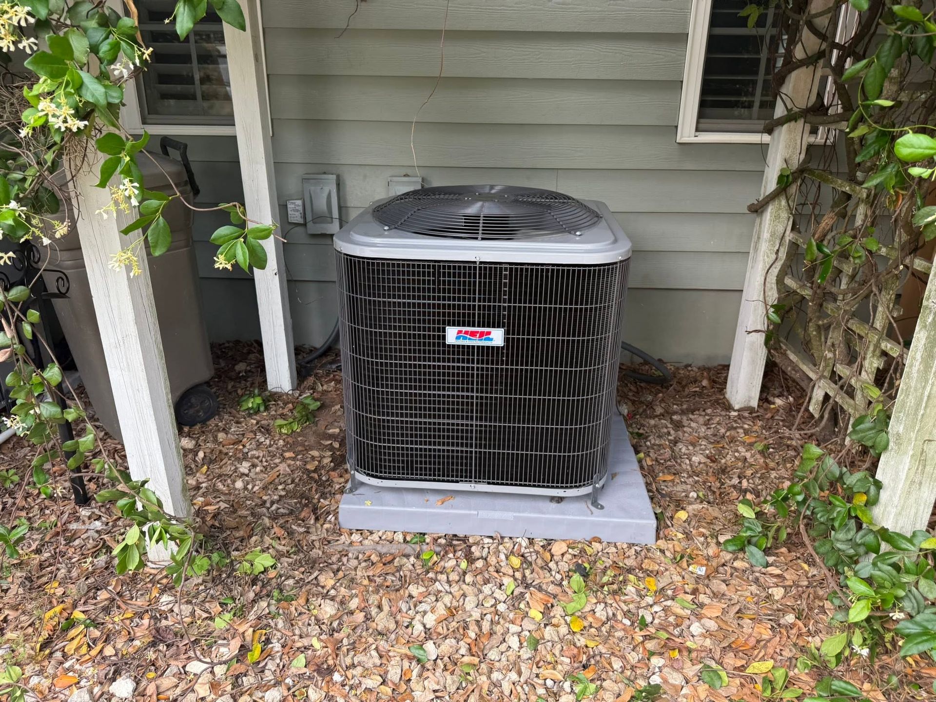 Outdoor air conditioner unit beside a house, partially enclosed by lattice and shrubs.