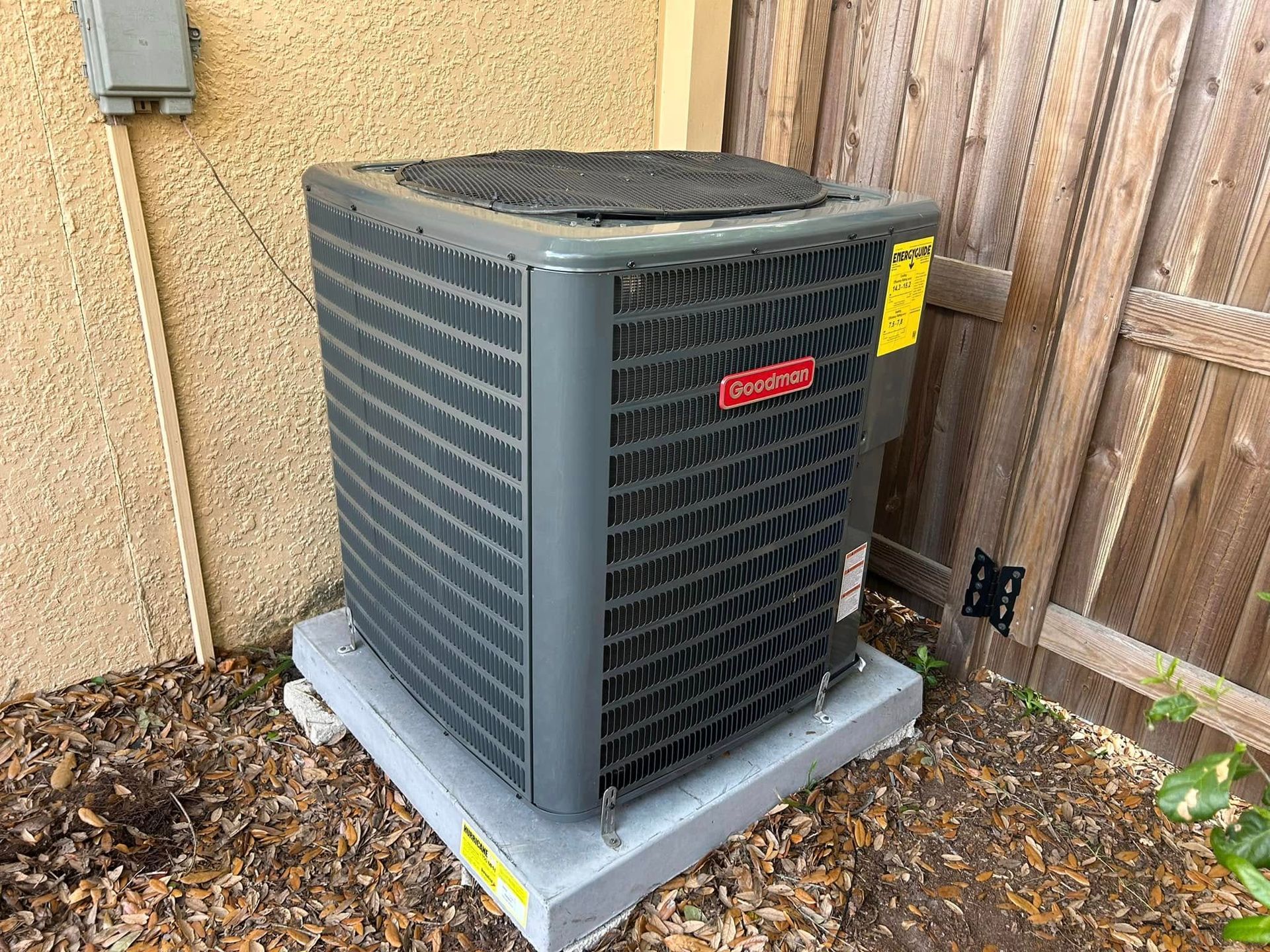 Outdoor HVAC condenser unit beside a house wall and wooden fence on a bed of fallen leaves