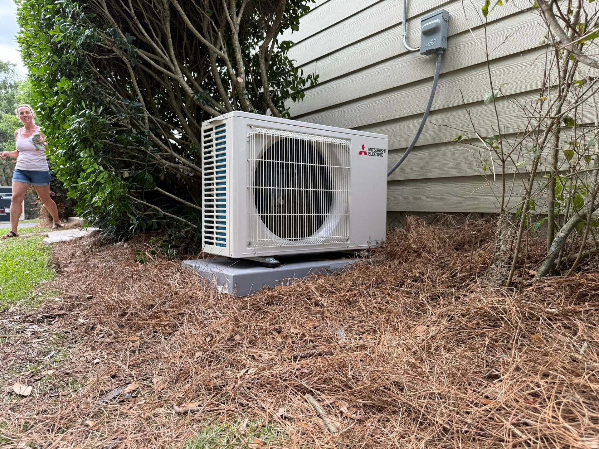Outdoor air conditioner unit beside a house, near shrubs and mulch.