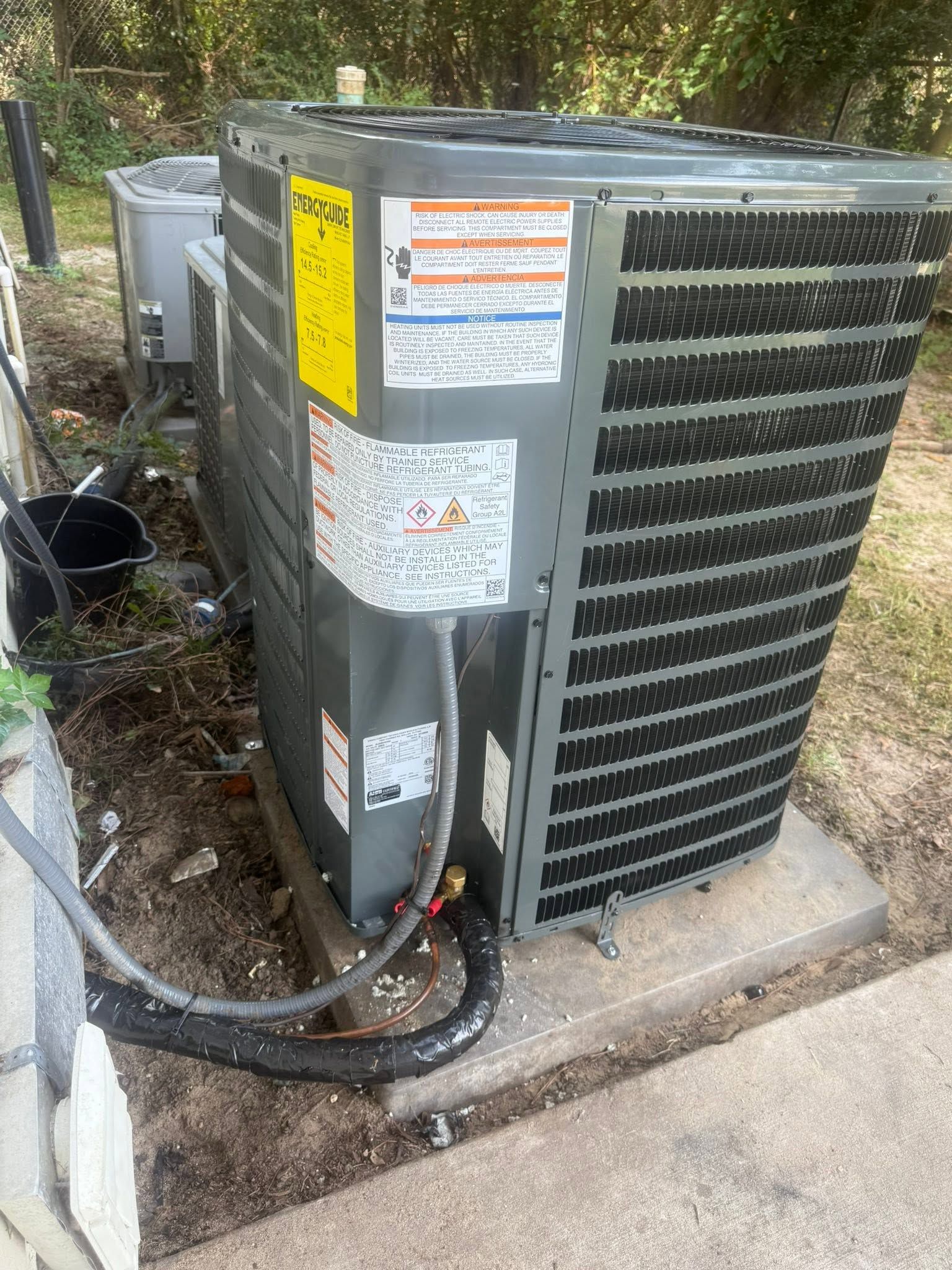 Outdoor air conditioning unit beside a sidewalk, with visible warning labels and connected hoses.