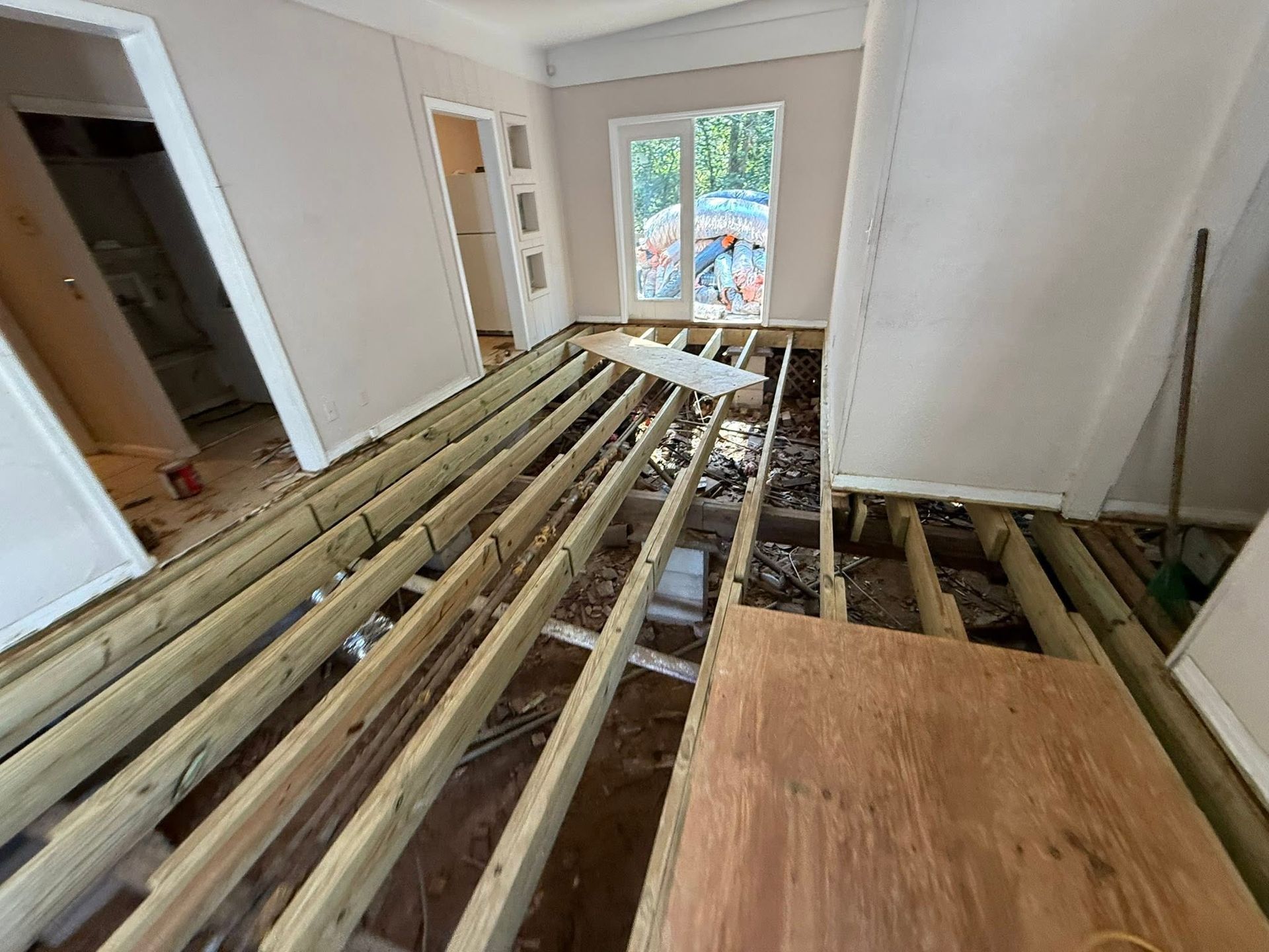 Interior room under renovation with exposed floor joists and plywood subfloor near a window.