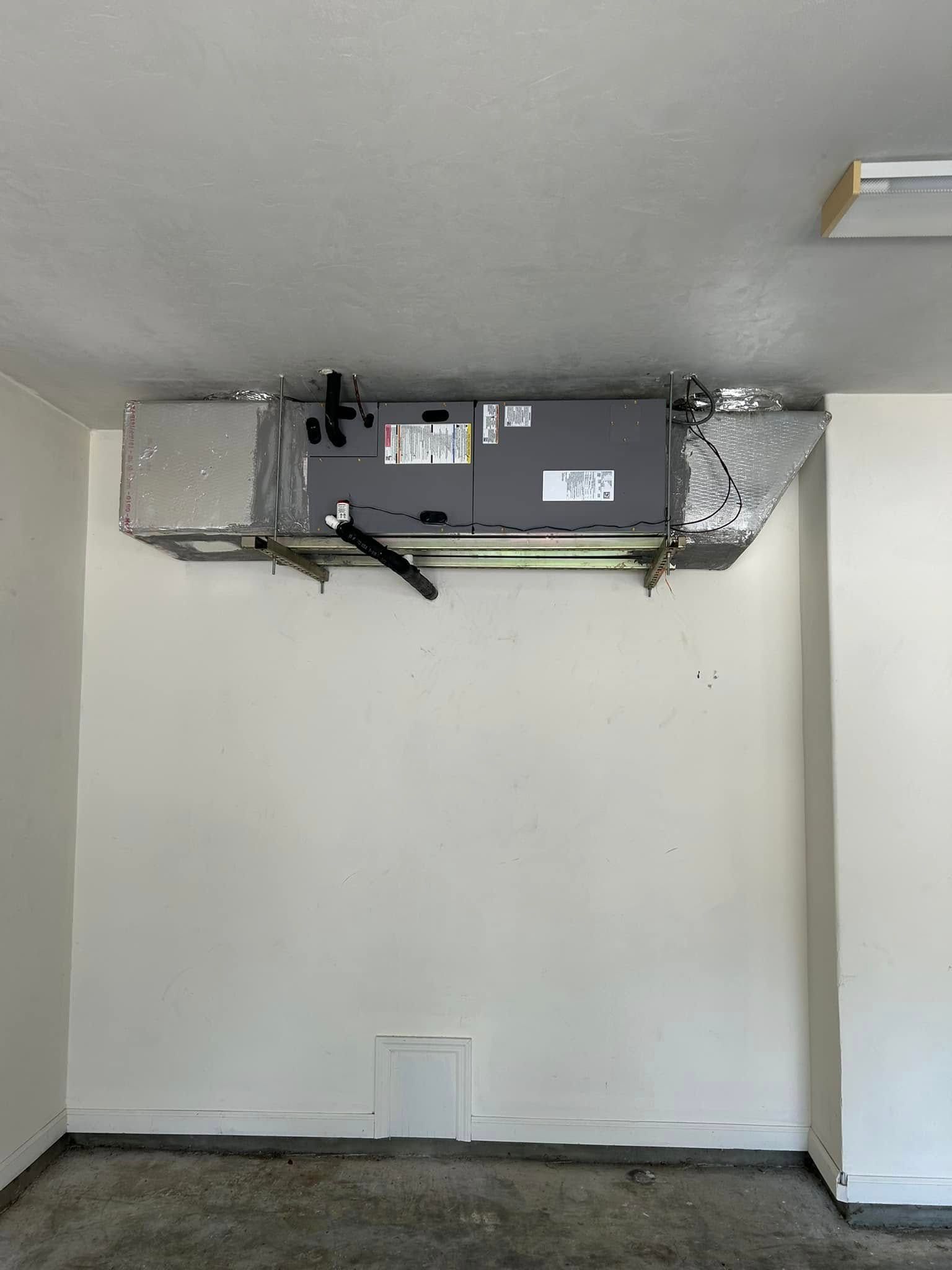 Wall-mounted HVAC unit in a plain white room with exposed ductwork near the ceiling