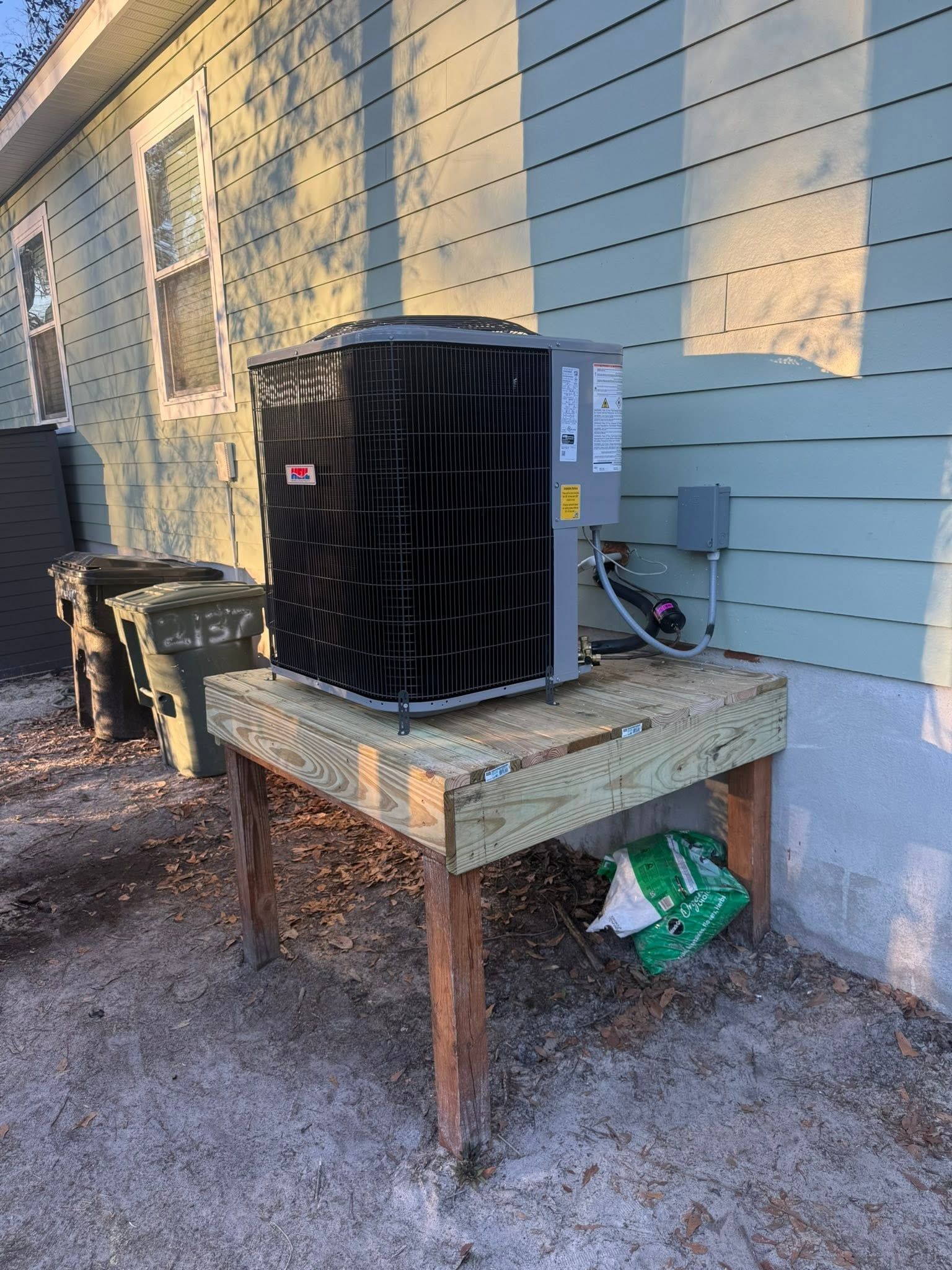 Outdoor HVAC unit on a wooden stand beside a house wall, with wires and a trash bag underneath.