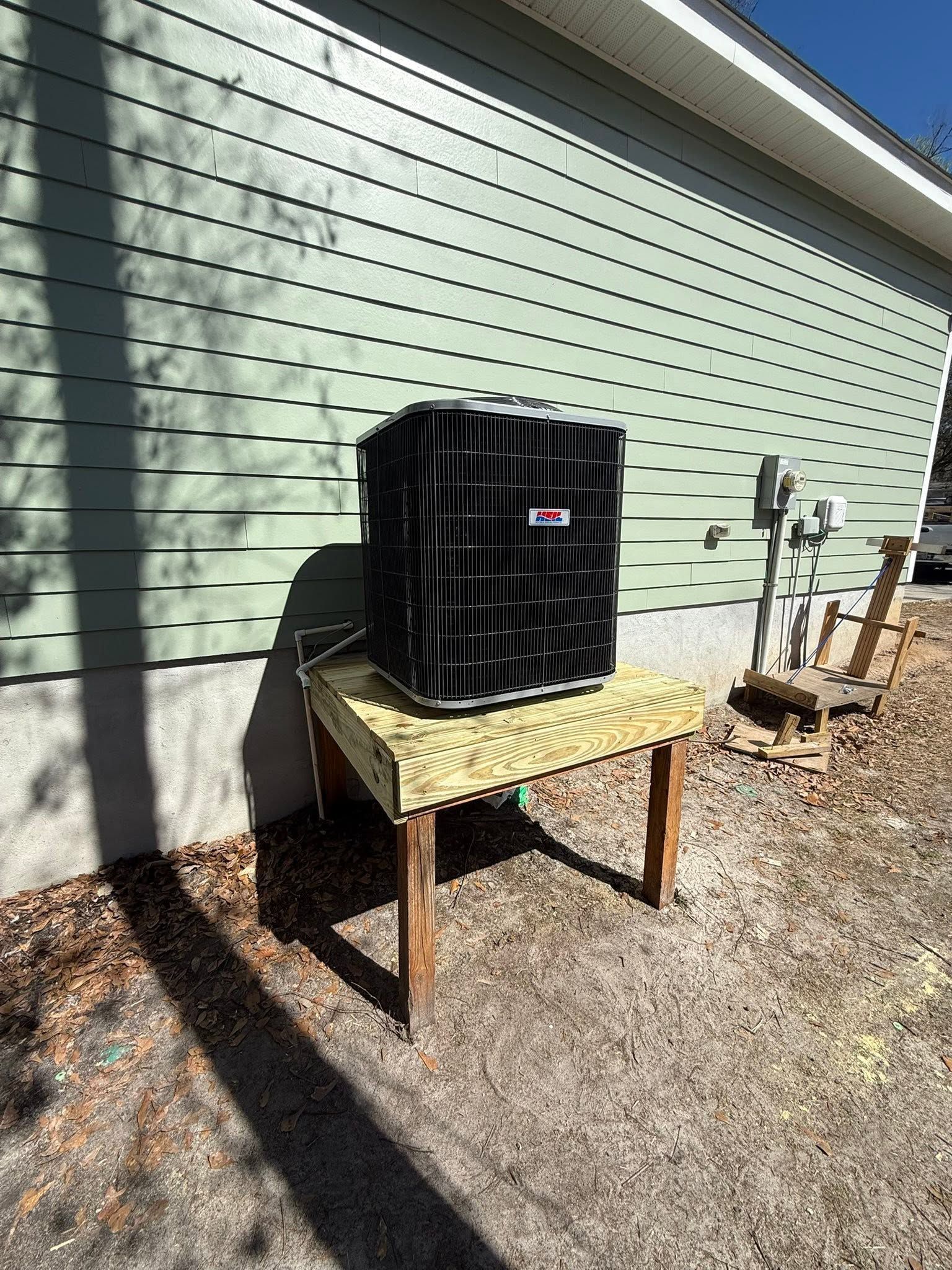 Black outdoor unit on a small table beside a house wall in a gravel yard, with plants nearby