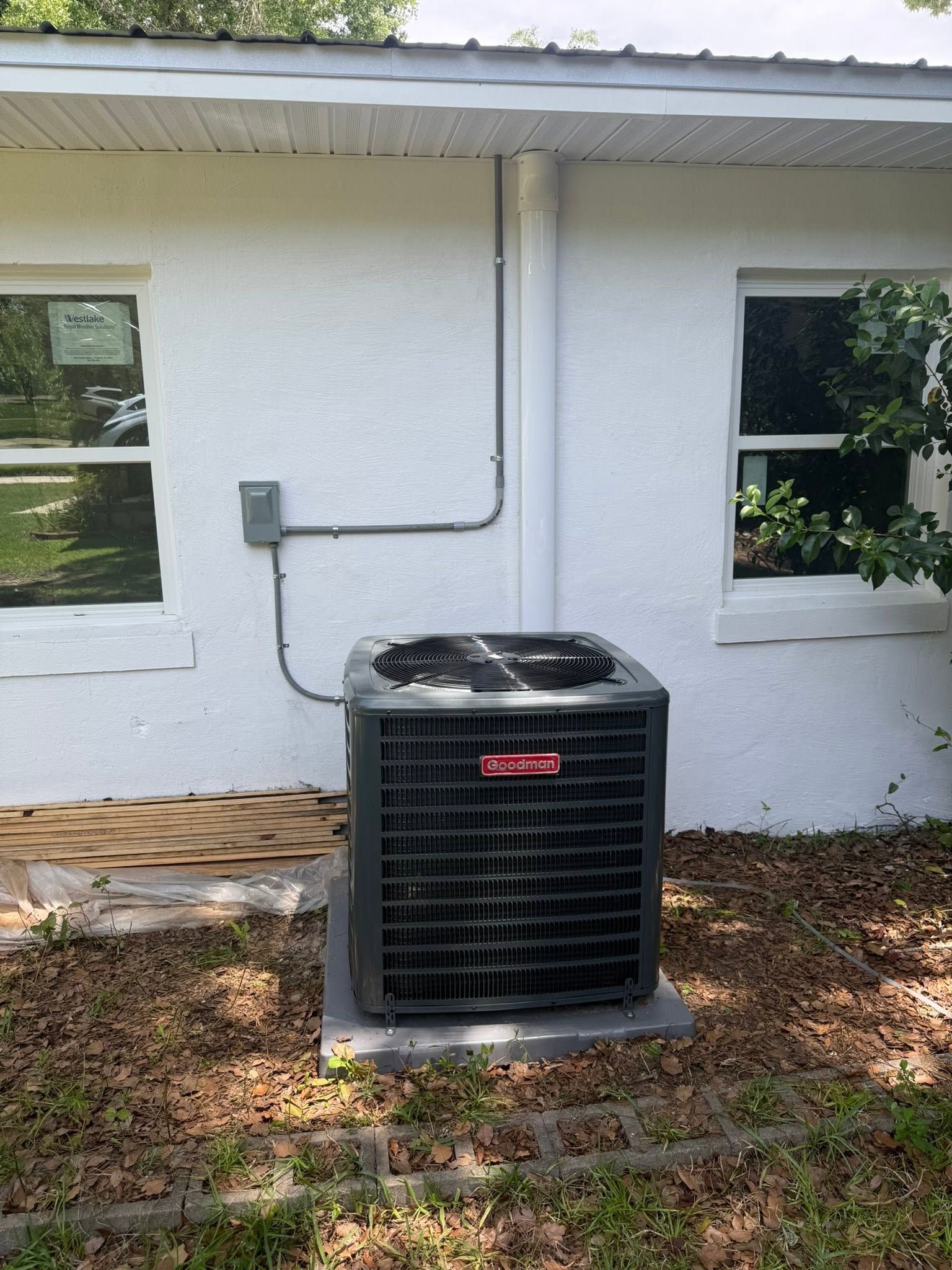 Outdoor air conditioner unit beside a white house wall with two windows and visible utility conduit