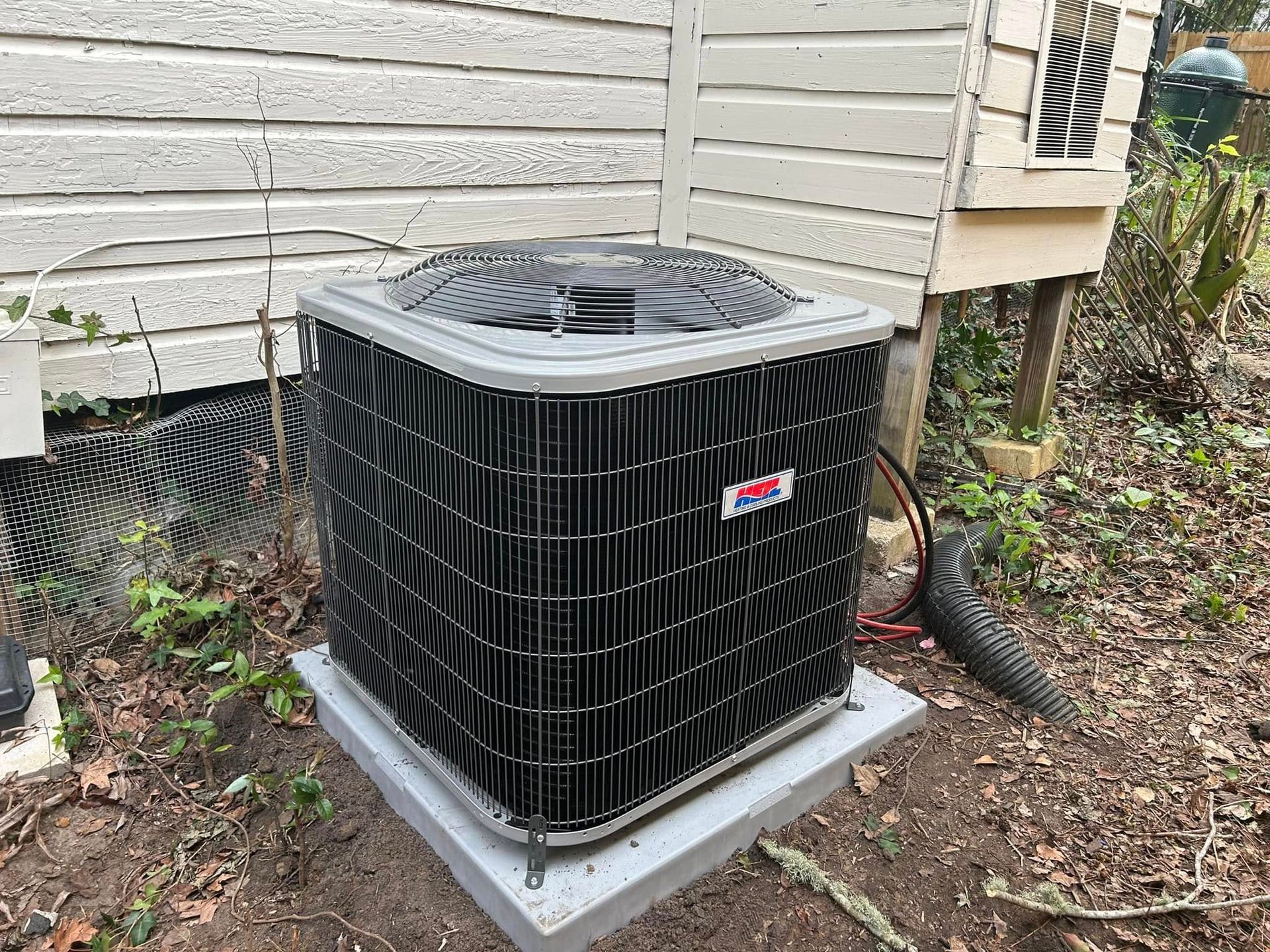 Outdoor air conditioner unit beside a house, with black grille and gray base on dirt and leaves
