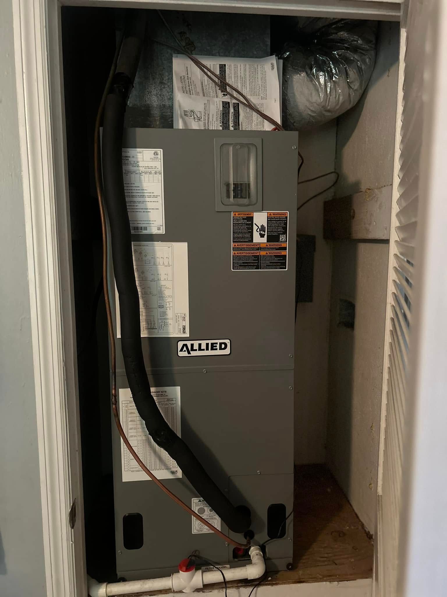 Gray HVAC unit in a narrow closet, with exposed pipes and wiring around it.