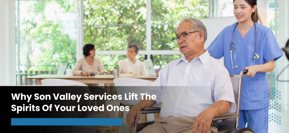 Caregiver in blue beside an older man in a wheelchair, with two people in a bright, sunlit room.