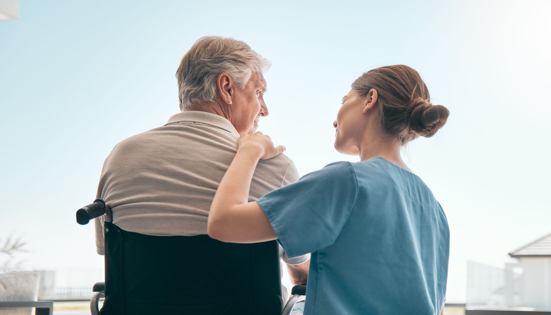 Caregiver in blue shirt speaking with an older person in a wheelchair, seen from behind in a bright room.