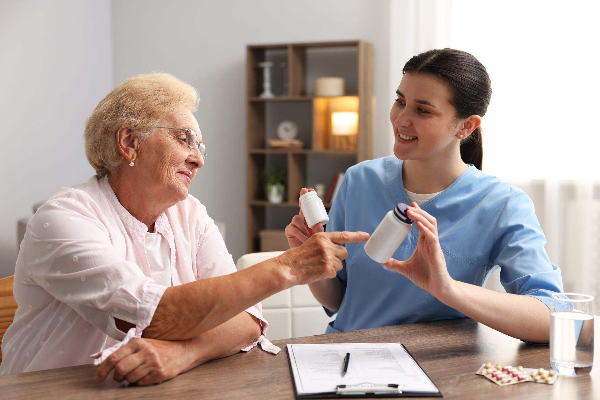 Caregiver handing medication to a smiling patient at a table with a clipboard and pills