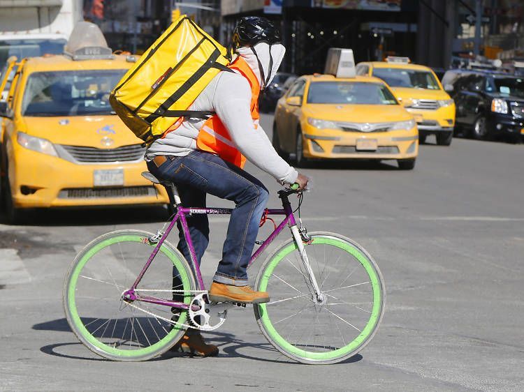 Cyclist in white and orange rides a purple bike with a yellow backpack past yellow taxis on a city street