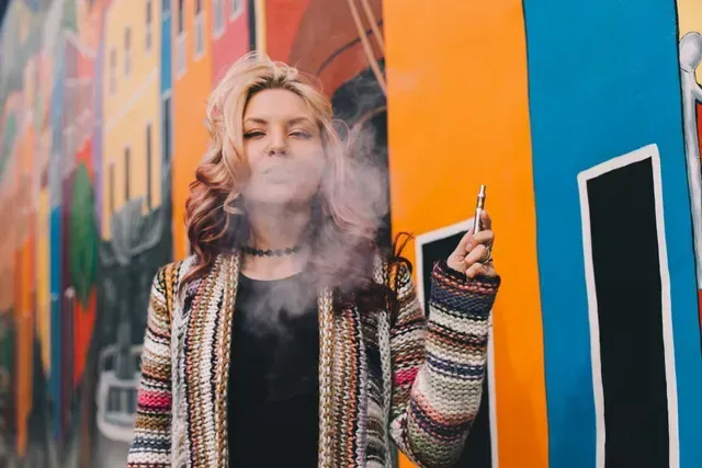 Person smoking beside a colorful mural, wearing a striped cardigan and black top.