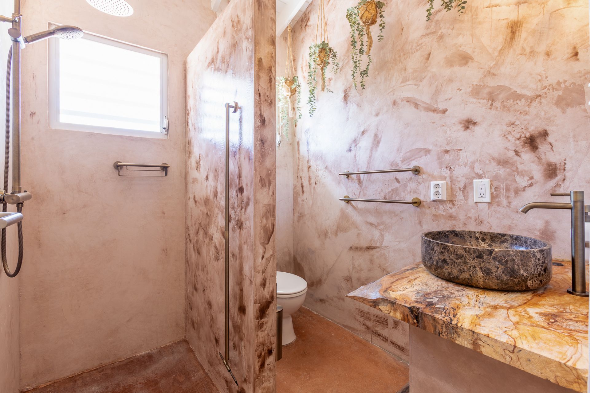 Rustieke badkamer met stenen wastafel, douche en toilet. Muren met structuurverf, raam en bloemaccenten.