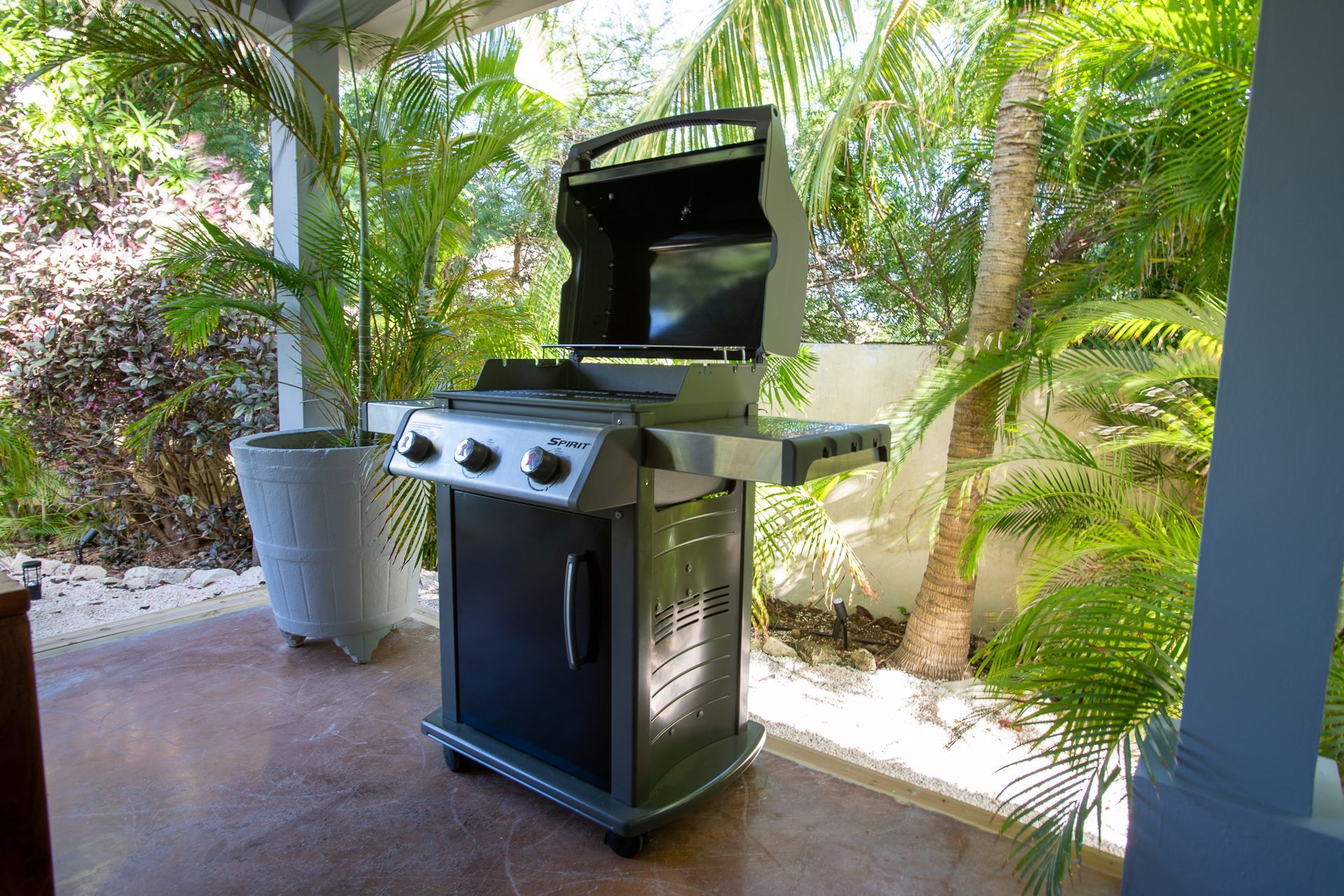 Een gasbarbecue op een terras, omgeven door weelderige groene planten en een witte pilaar.