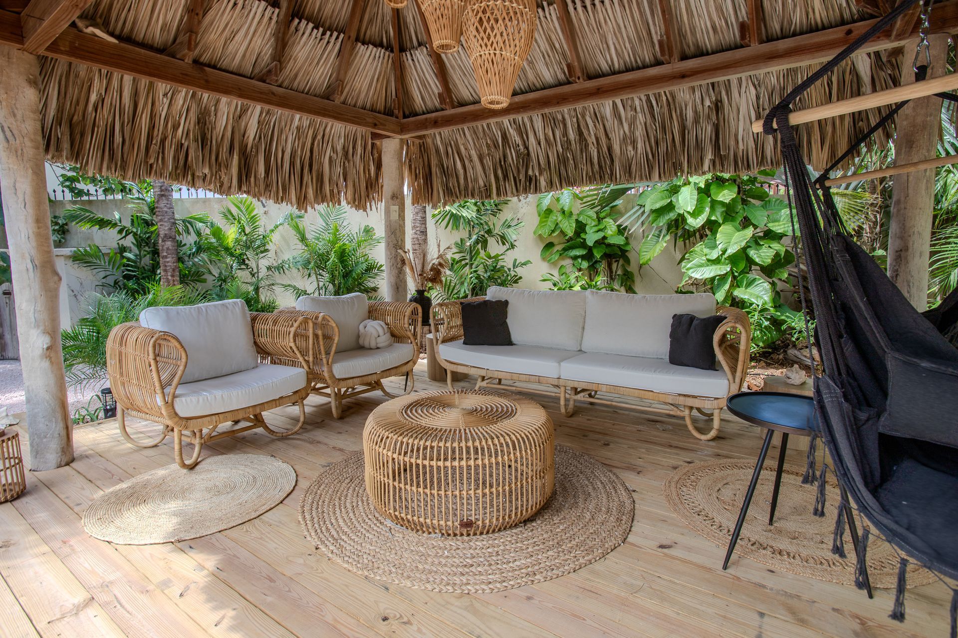 Een zitgedeelte buiten onder een rieten dak. Rotan meubilair, waaronder een bank, stoelen en een tafel.