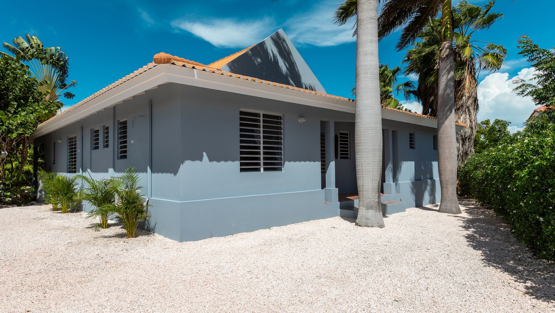 Grijs huis met wit grind, palmbomen en een blauwe lucht.