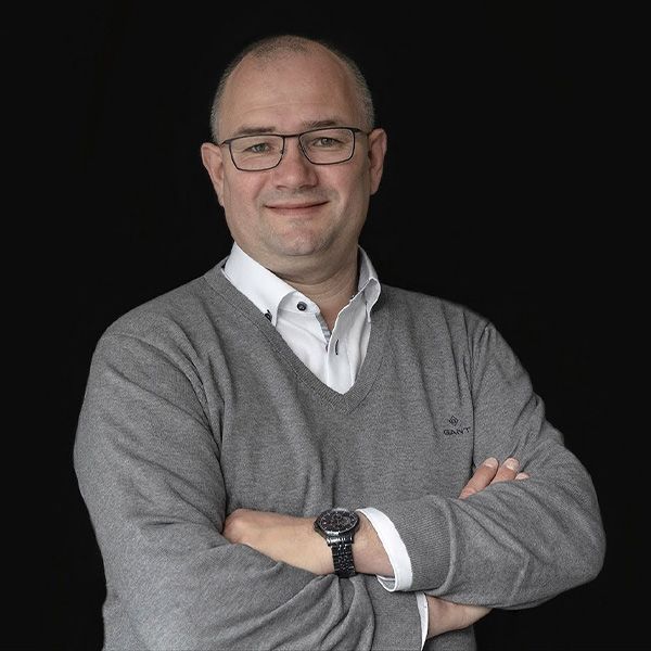 Jean-Pierre met een bril en een grijze trui staat met zijn armen over elkaar.