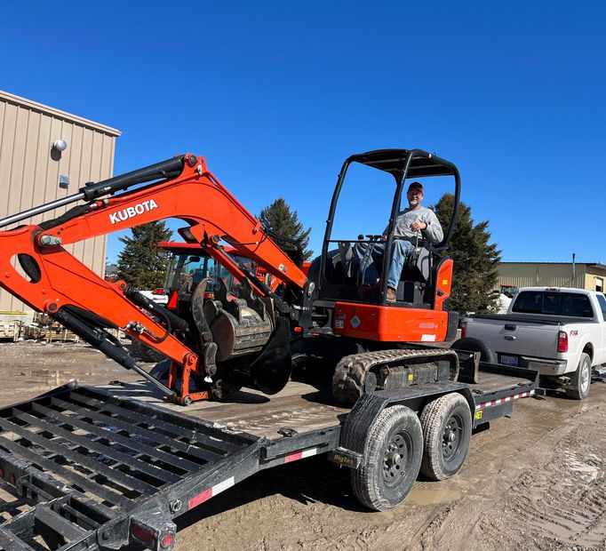 Orange mini excavator on a trailer at a construction site, with a worker in the cab and a pickup nearby.
