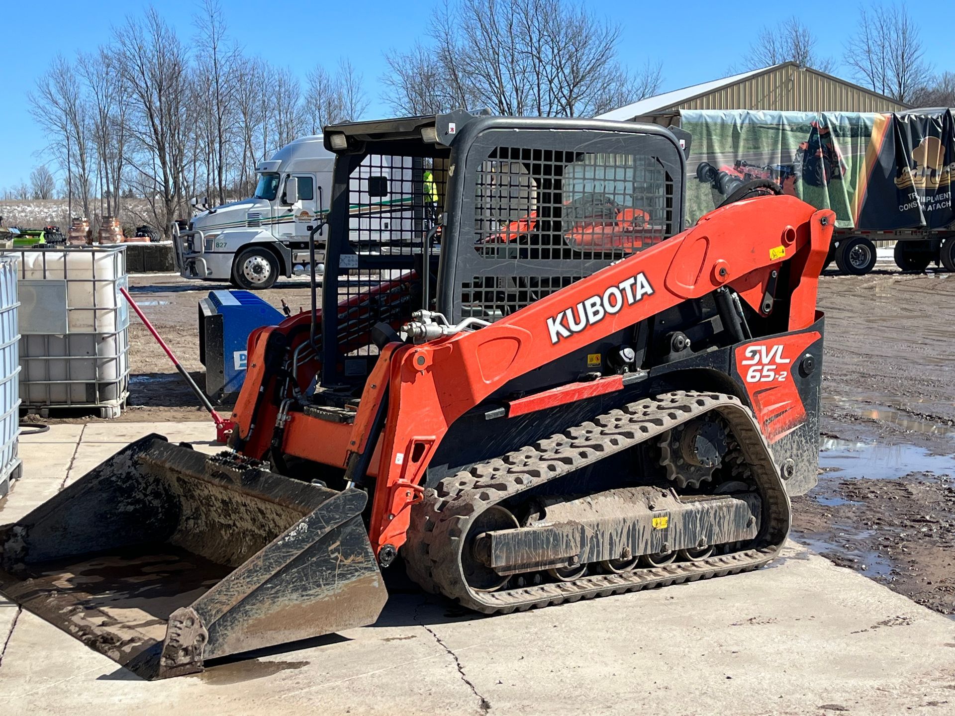 Orange Kubota skid steer with bucket on a concrete lot, pushing soil beside a white truck and shed.