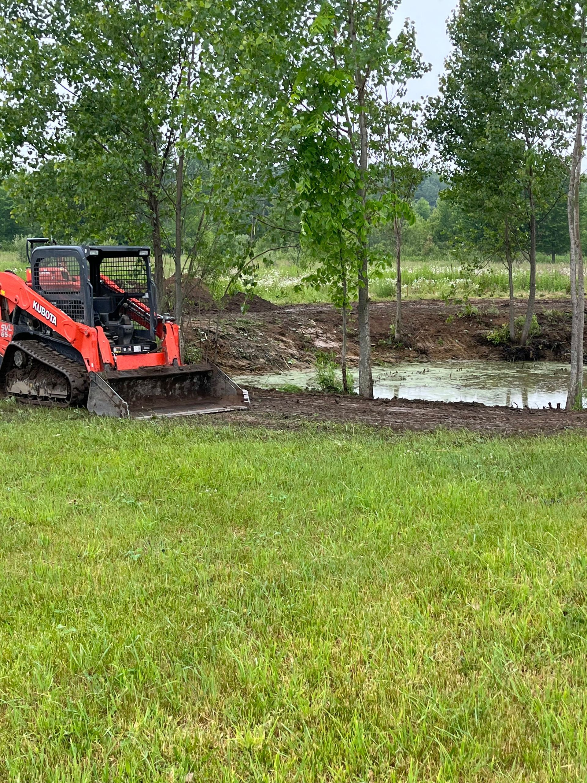 Orange skid steer near muddy pond beside green trees and grass