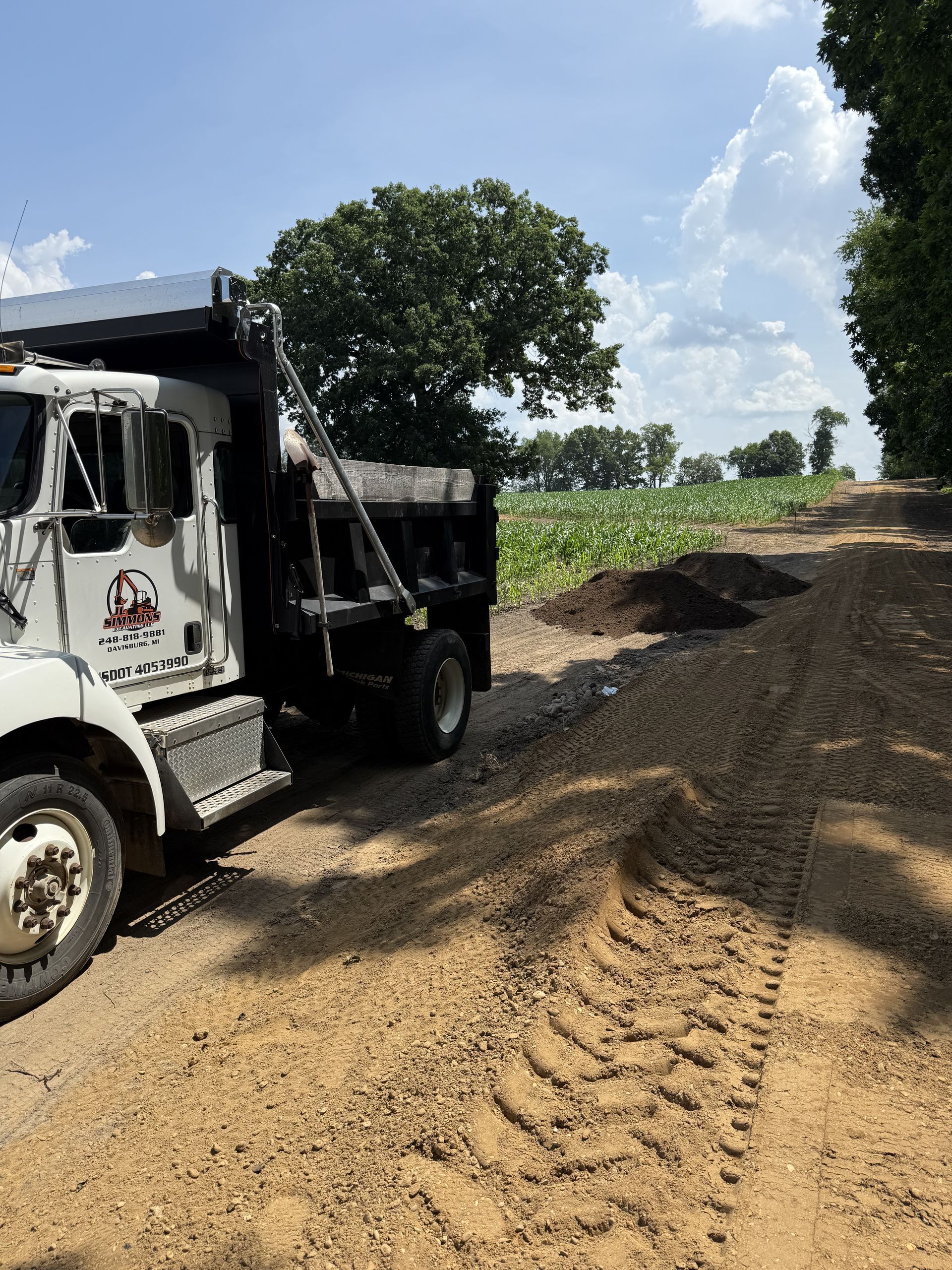 White dump truck on a dirt road beside a trench, with a green field and trees under a cloudy sky.