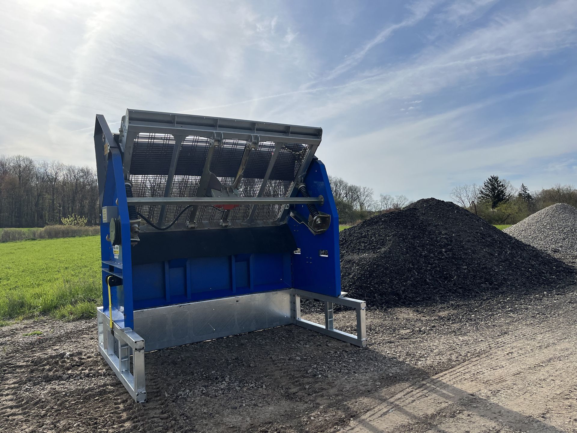 Blue industrial sorting bucket on gravel beside piles of rock under a cloudy sky
