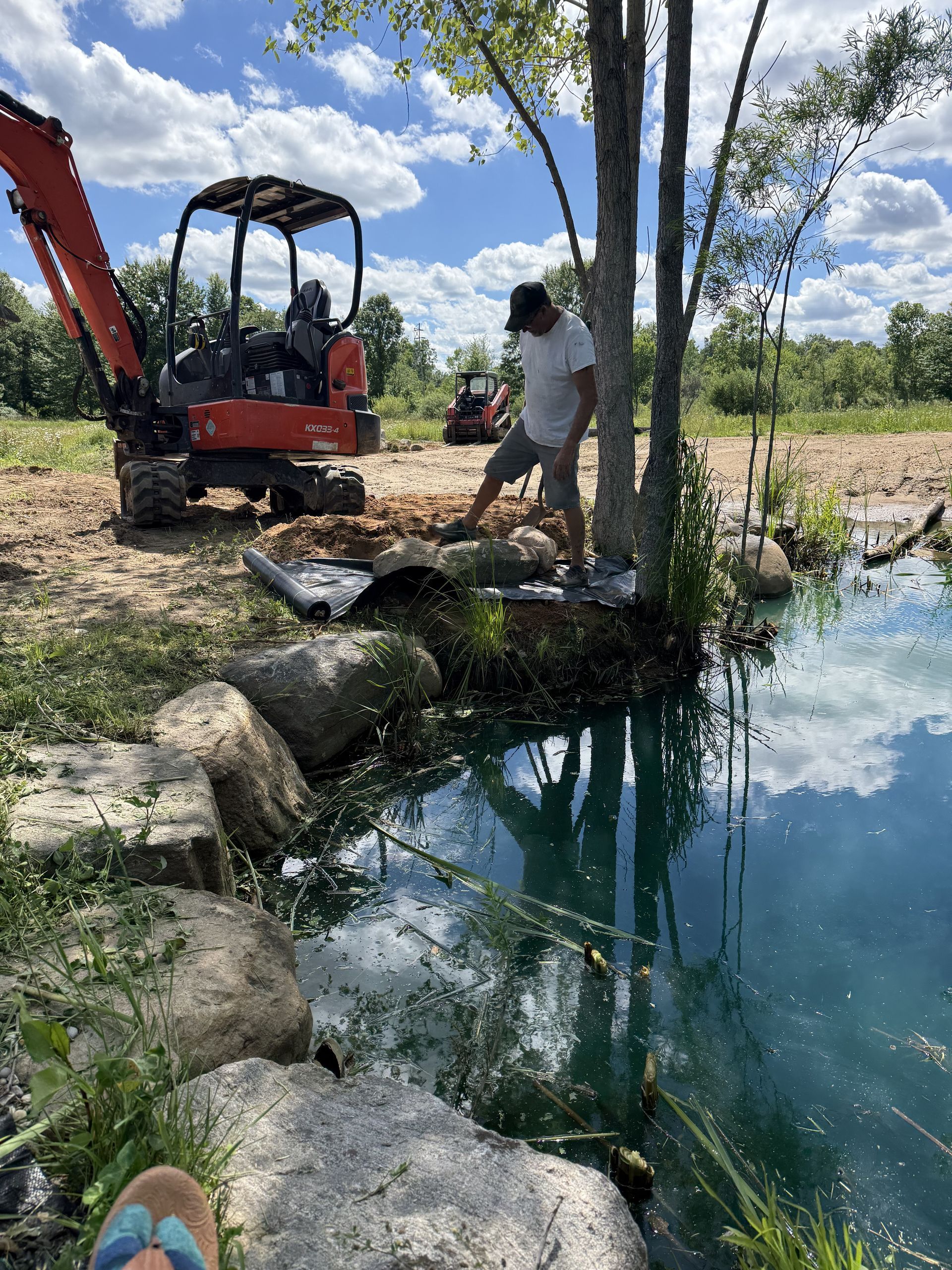 Excavator and worker beside a small canal, clearing debris in a grassy rural area.