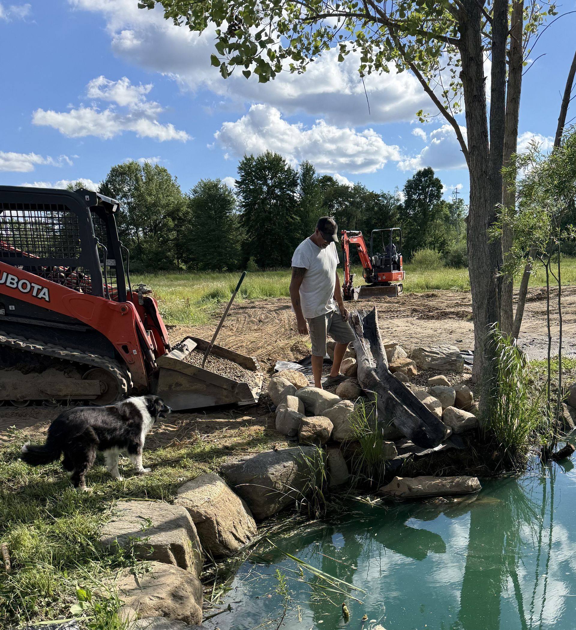 Workers and a skid steer clear debris beside a pond under a bright sky