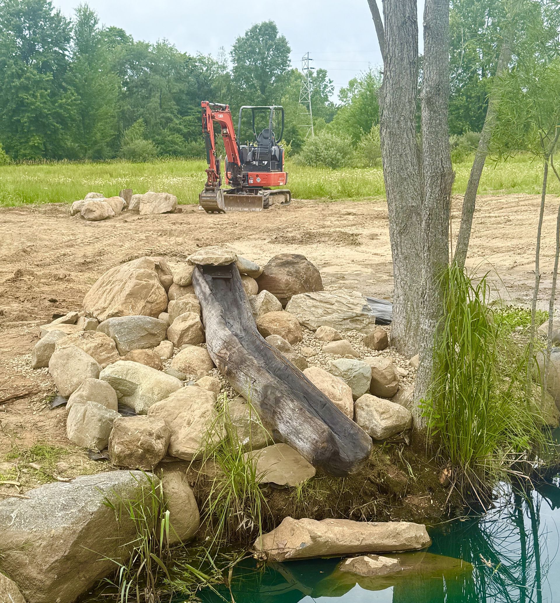 Small excavator on a rocky dirt bank by a stream, with a log or pipe leading into the water.