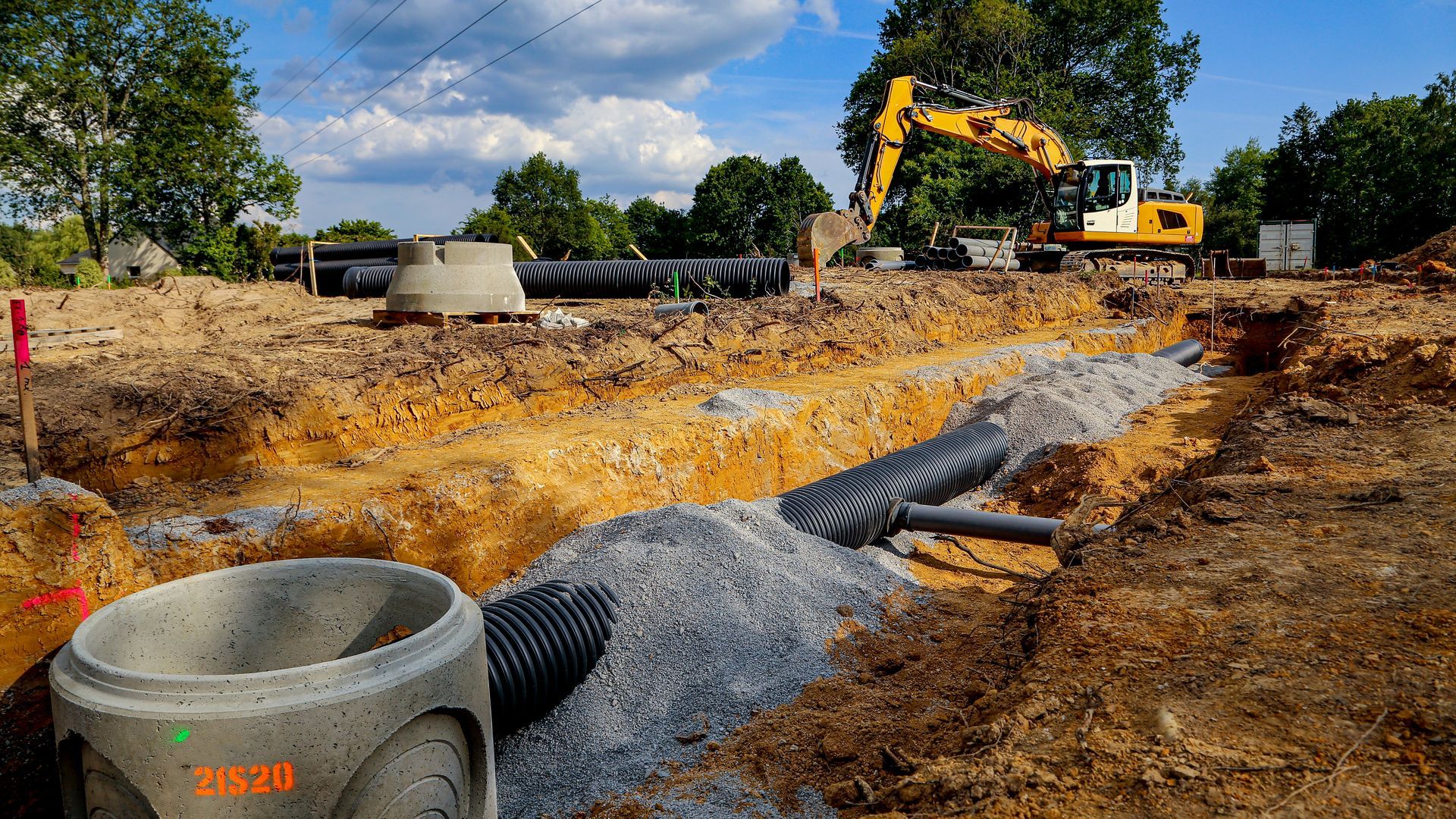 Construction site with excavator, gravel piles, and drainage pipes under a cloudy sky