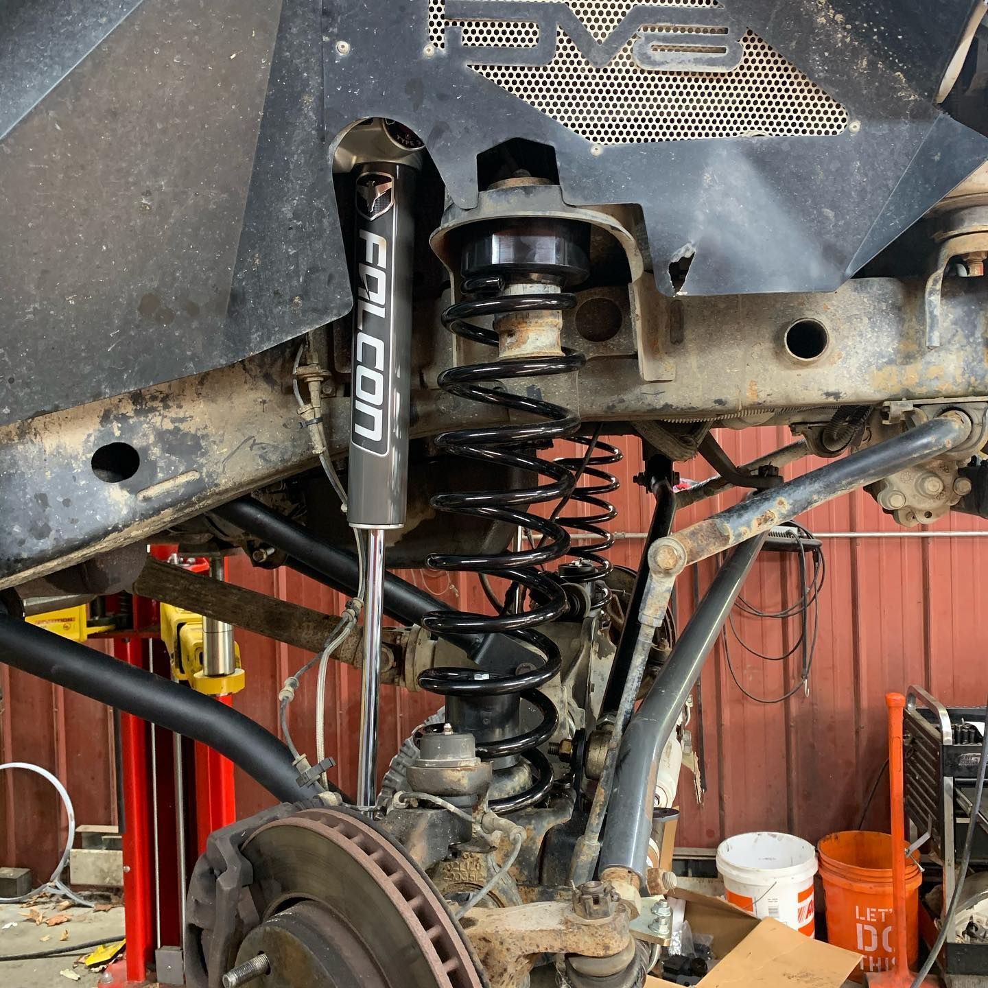 Jeep suspension components: shock absorber, coil spring, control arm, brake rotor, and a metal shield in a garage setting. Jeep suspension components: shock absorber, coil spring, control arm, brake rotor, and a metal shield in a garage setting.