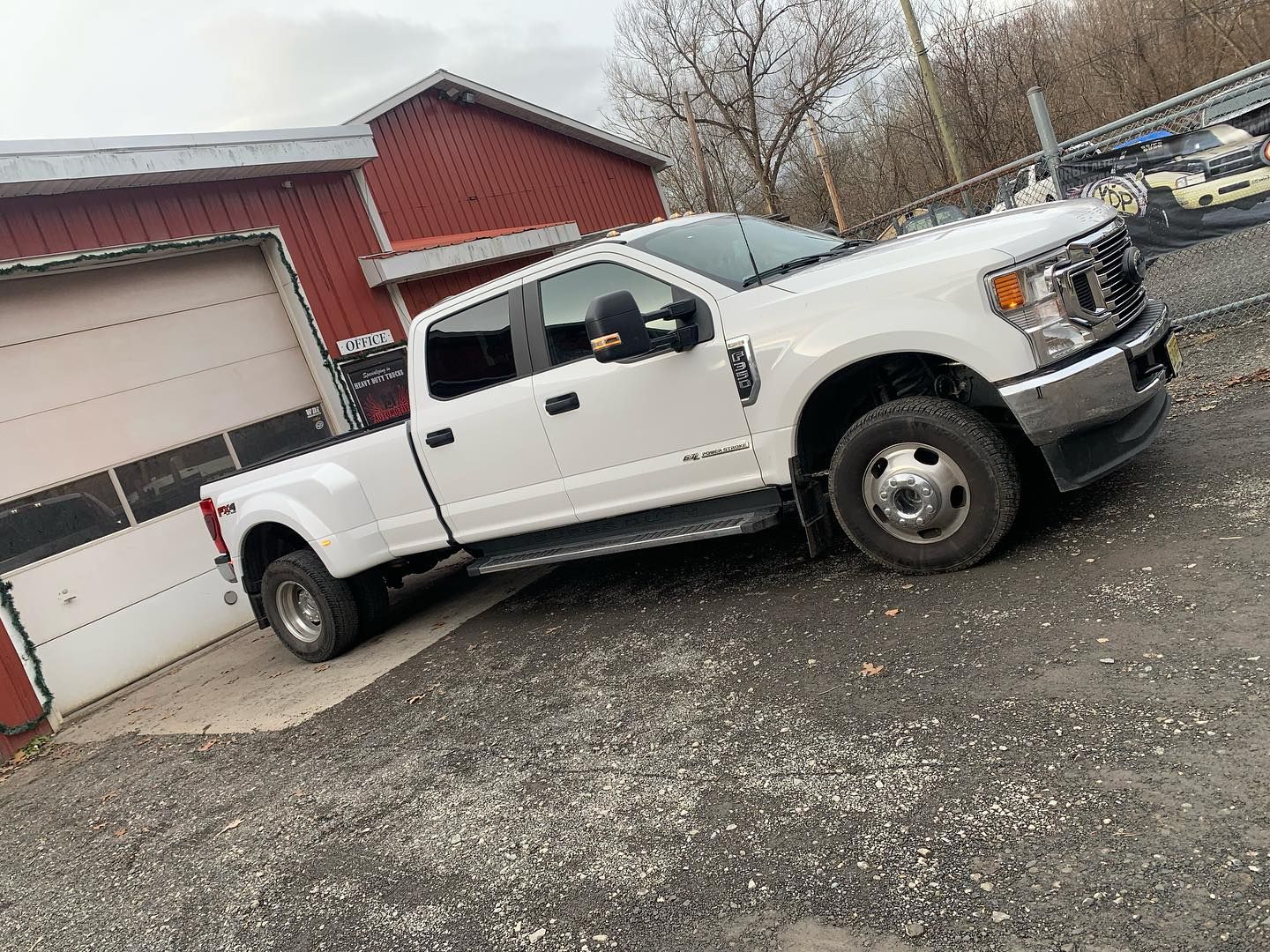 White Ford F-350 dually pickup truck parked next to a red building with a garage door, gravel driveway.