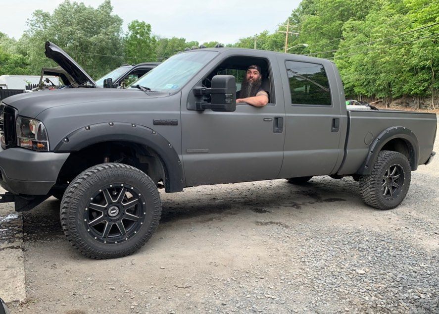Man in gray pickup truck smiles at camera. Gray truck has black wheels and fender flares, parked outdoors. Man in gray pickup truck smiles at camera. Gray truck has black wheels and fender flares, parked outdoors.