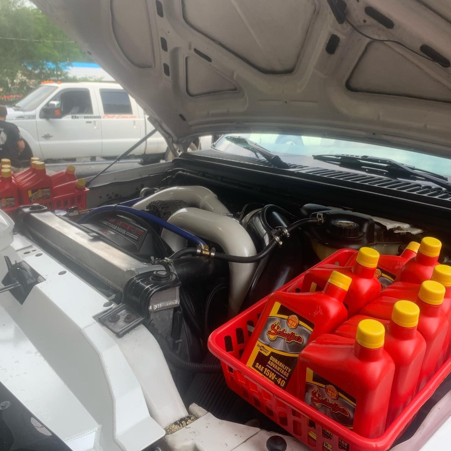 Open hood of a white truck. A basket of red oil bottles sits beside the engine. Another truck is in the background. Open hood of a white truck. A basket of red oil bottles sits beside the engine. Another truck is in the background.