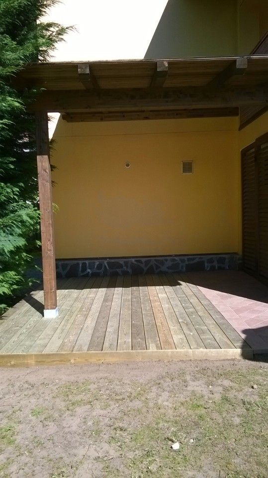 Veranda coperta in legno con parete gialla, base in pietra e prato antistante.
