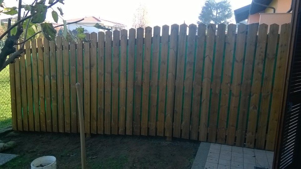 Recinzione in legno nel cortile sul retro con doghe verticali, sole e un piccolo patio pavimentato sulla destra.