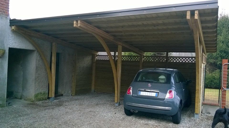 Auto blu parcheggiata sotto una tettoia di legno accanto a un vialetto di ghiaia e a un muro intonacato.