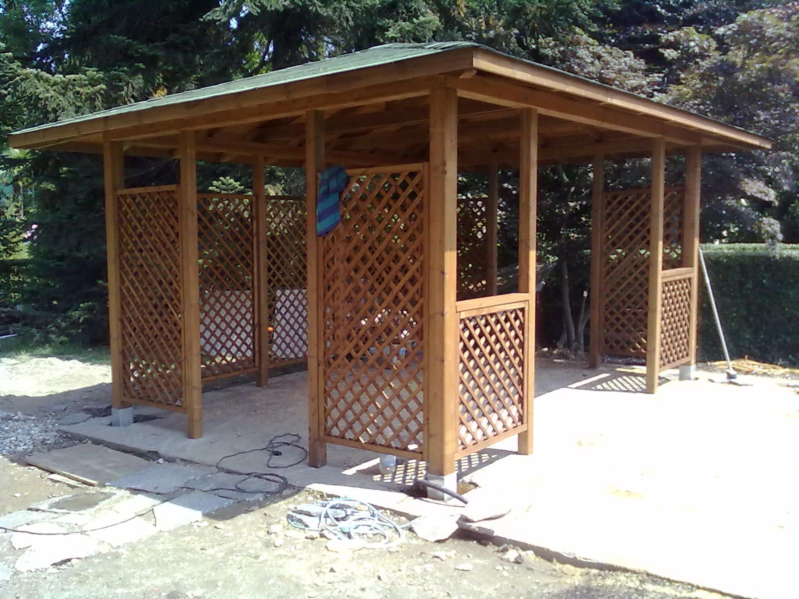Gazebo da esterno in legno con pareti a traliccio e tetto spiovente su un patio in pietra in una zona boschiva.