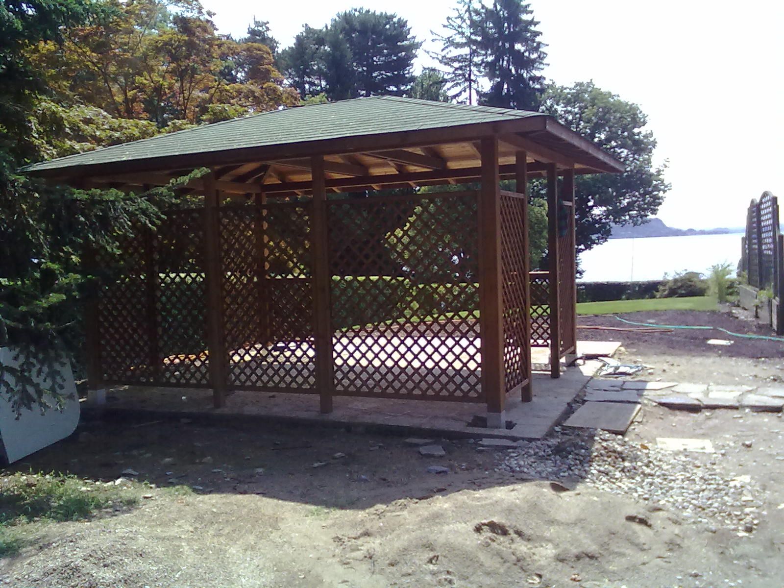 Gazebo in legno con traliccio e tetto verde in un cortile esterno soleggiato, circondato da alberi e terreno sterrato.