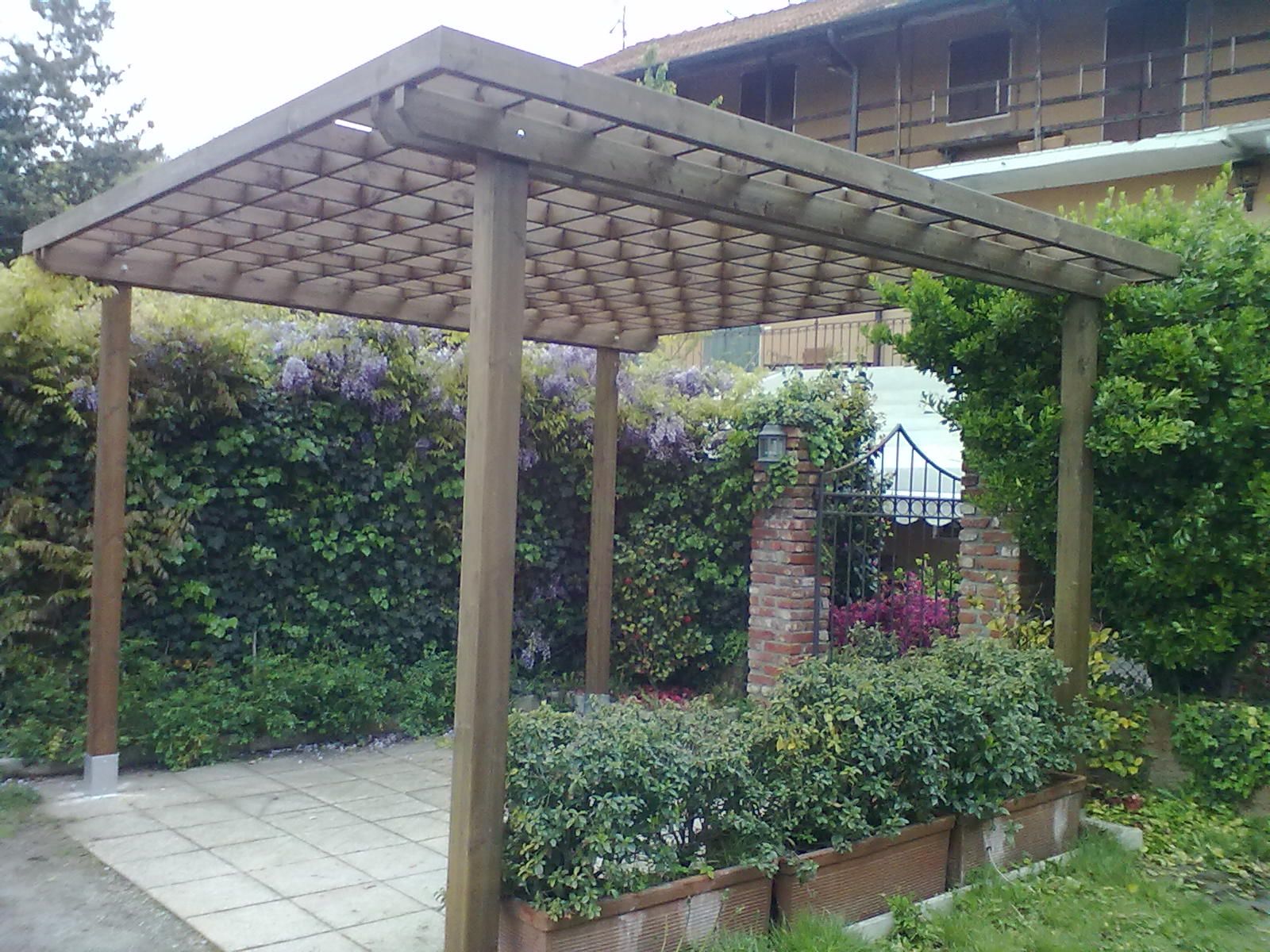 Patio in giardino con pergolato in legno, sentiero piastrellato e rigogliosi arbusti verdi accanto a una siepe e alla casa.