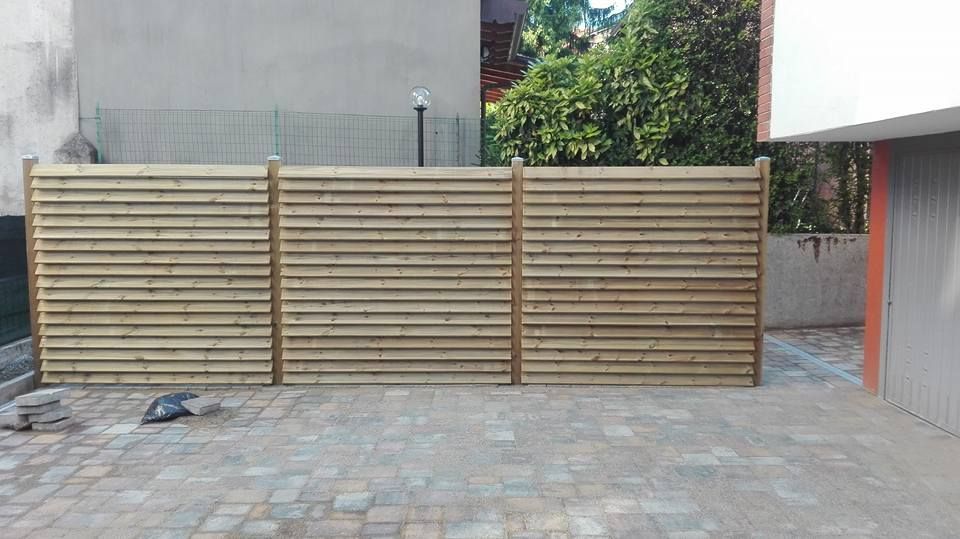 Una recinzione a doghe di legno attraversa un vialetto pavimentato accanto a un edificio grigio e a una porta del garage.