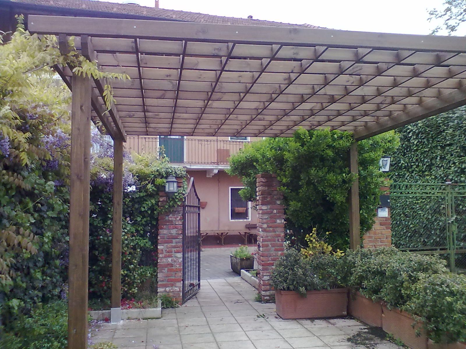 Un sentiero nel giardino, sotto un pergolato di legno, conduce all'ingresso di una casa in mattoni.