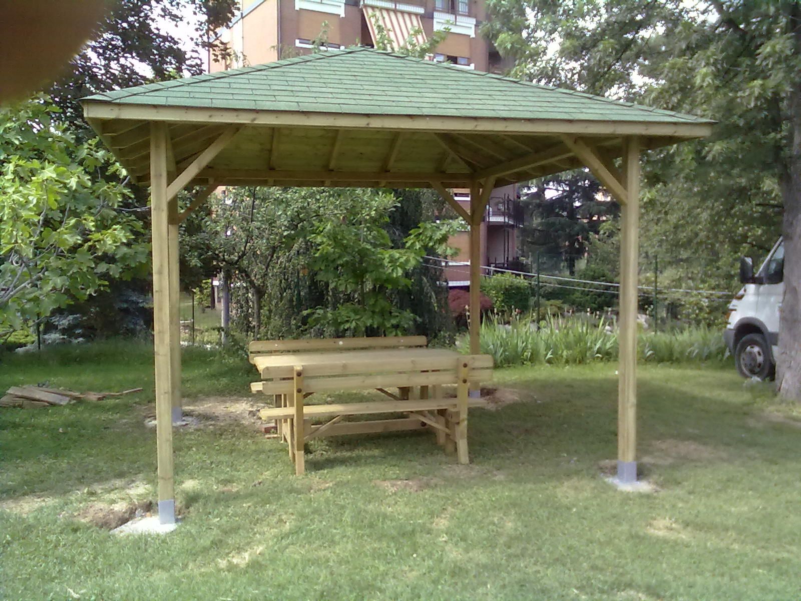 Gazebo in legno con tavolo da picnic in un prato, circondato da alberi e arbusti.