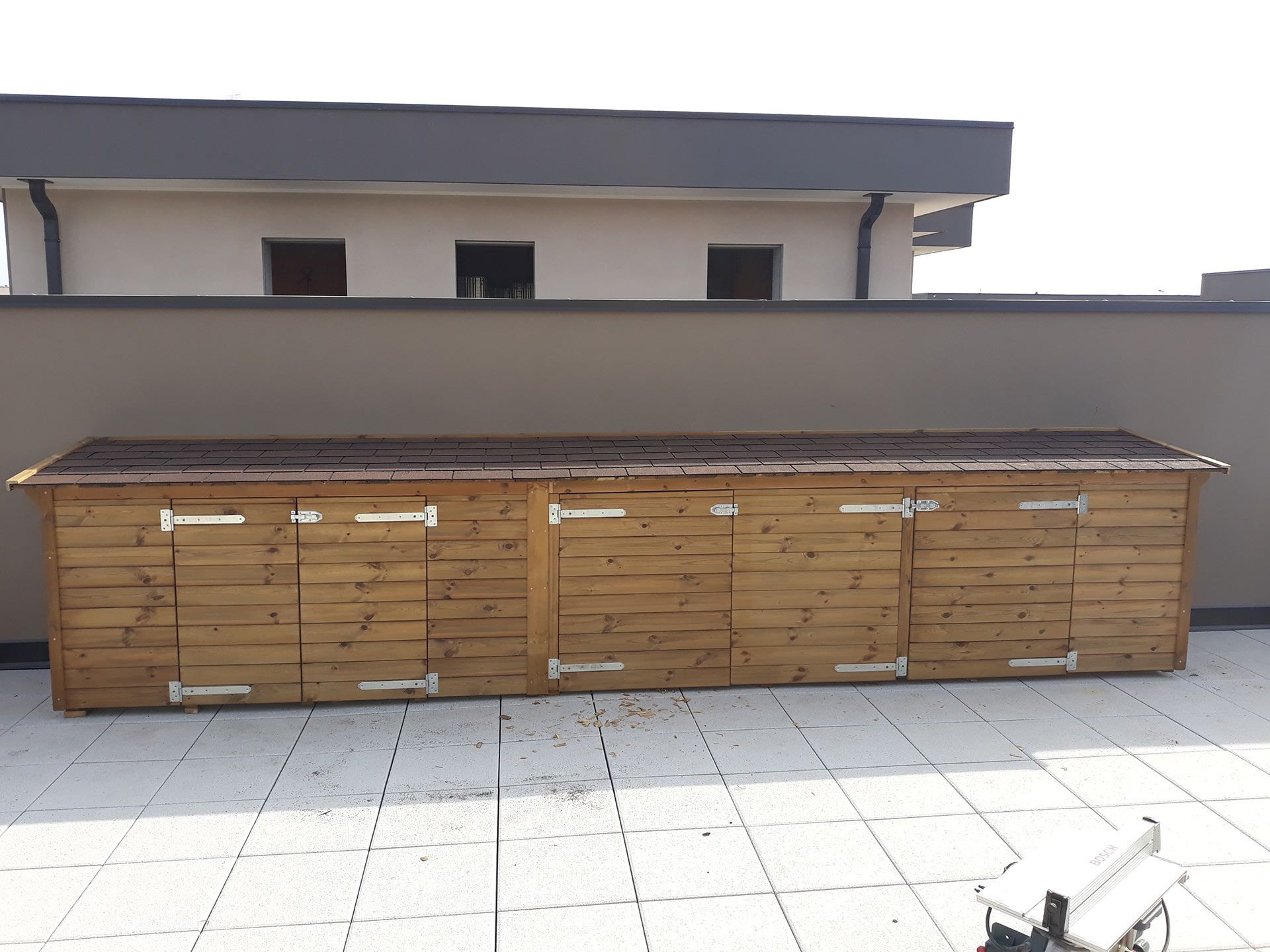 Panche contenitore in legno per esterni lungo la parete di una terrazza sul tetto, con pavimento piastrellato e una sedia bianca davanti.