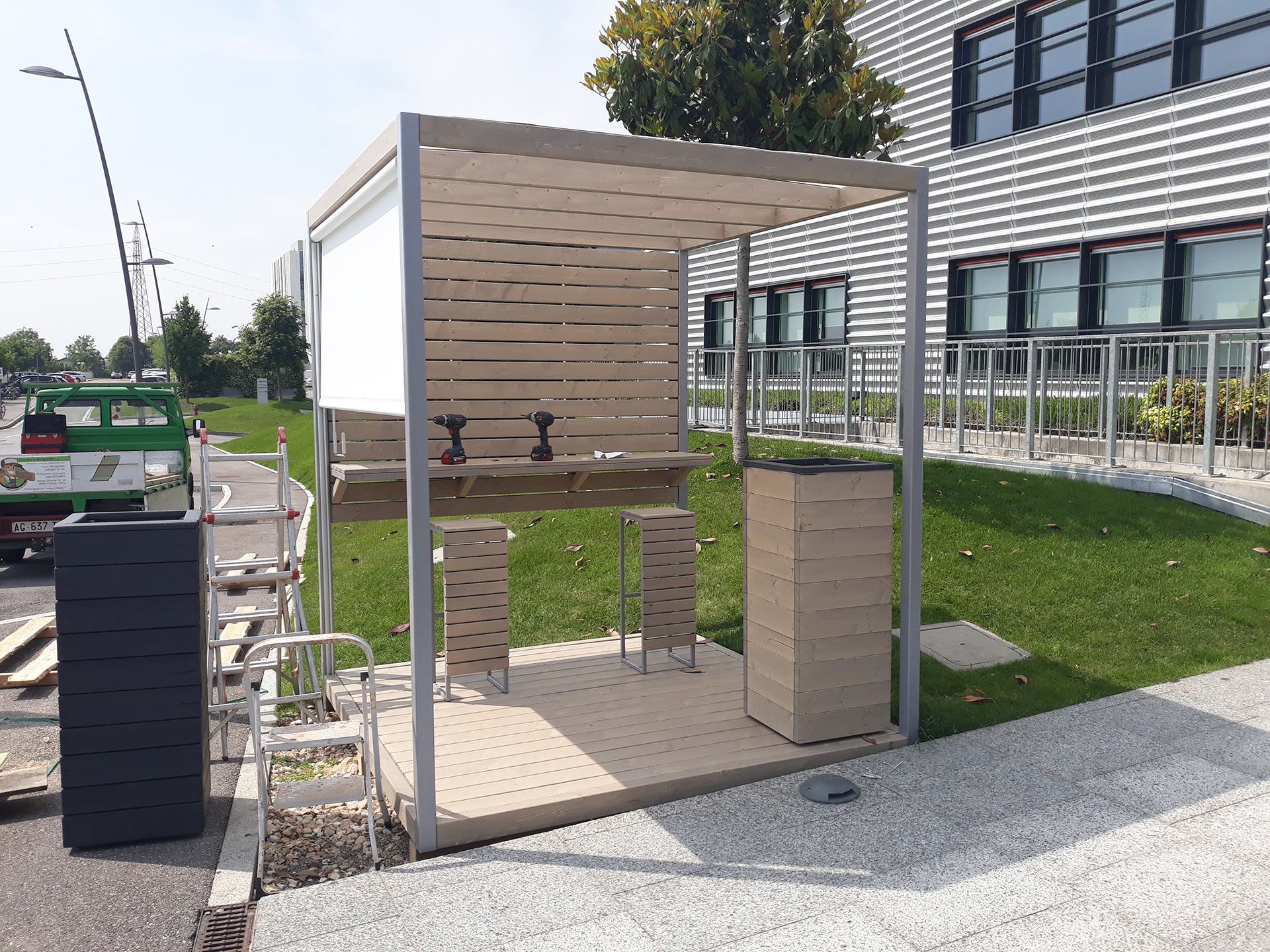 Chiosco o cabina di accessibilità in legno per esterni, con rampa, situato accanto a un edificio moderno su un sentiero di ghiaia.