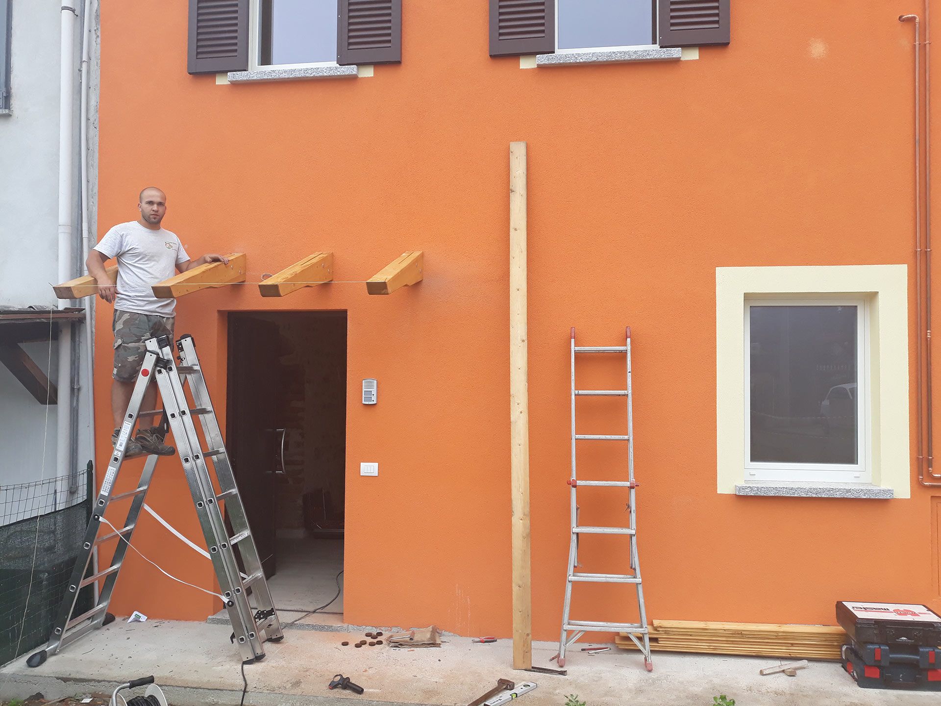 Un operaio su una scala dipinge l'esterno di una casa arancione con due scale e una finestra.