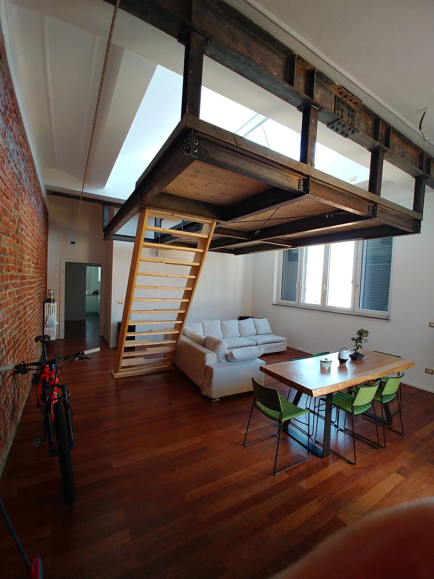 Camera da letto soppalcata con scala, divano bianco, tavolo e pavimento in legno sotto una piattaforma letto rialzata.