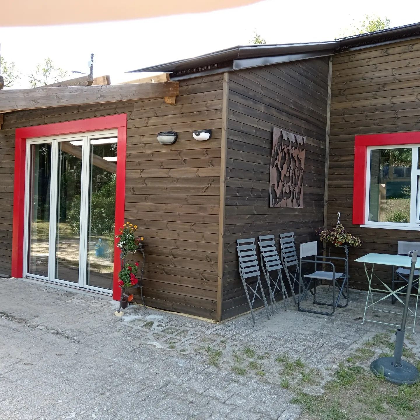 Esterno di un caffè in legno con finestre rifinite di rosso, tavolini all'aperto e sedie impilate accanto a un sentiero di ghiaia.